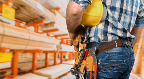 Construction tradesman holding hardhat under arm and looking at stacked lumber
