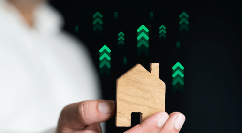 Hand holding wooden house with upward green arrows to indicate increased green building practices